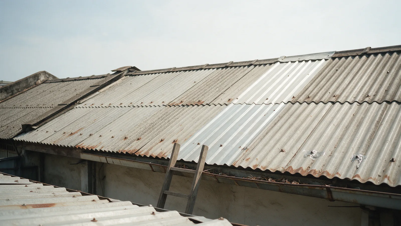 Illustrative ageing corrugated metal roof on an older shoplot row, rusting at the fastener lines, with one newer sheet and a ladder at the gutter