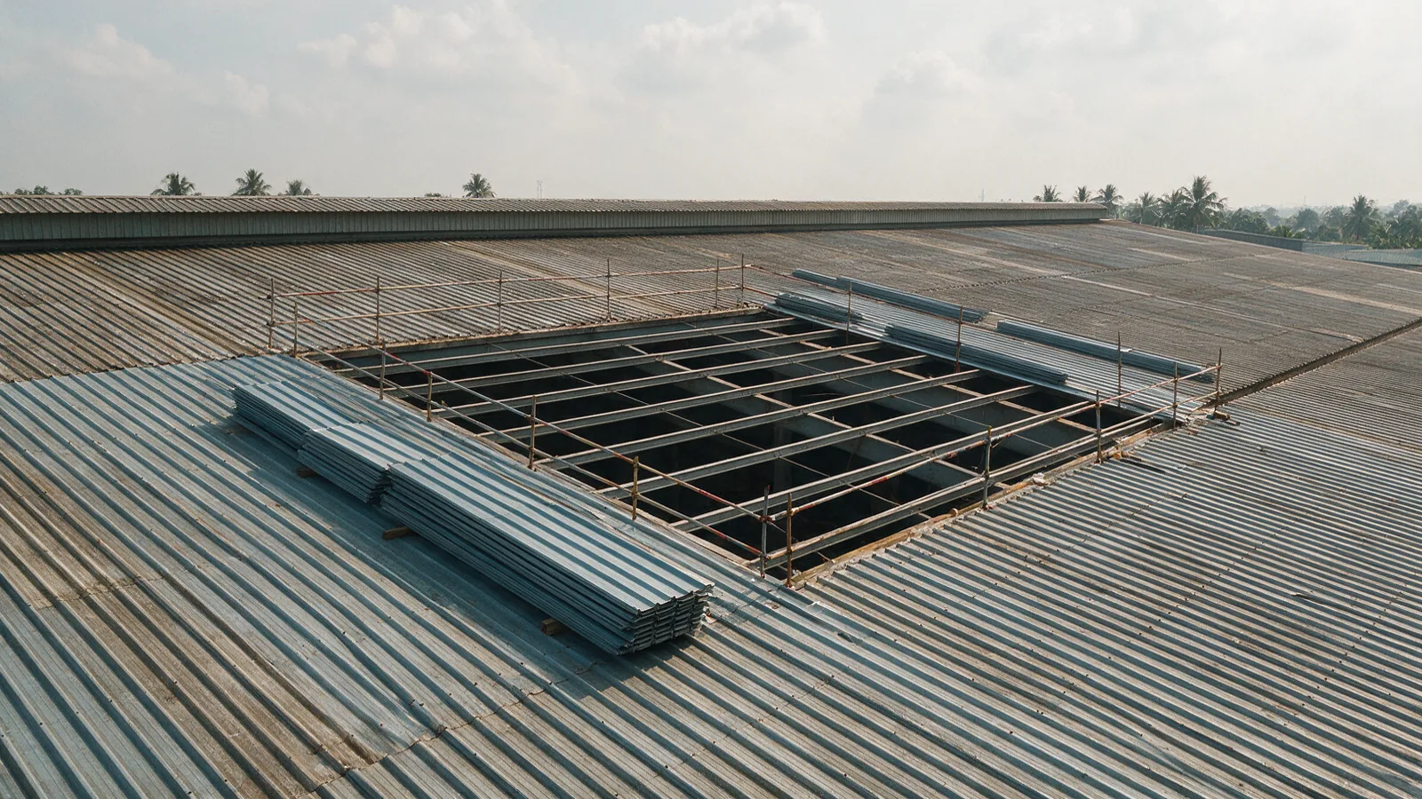 Illustrative view of a large metal roof part-way through reroofing, with one bay stripped to the purlins, new sheets stacked alongside and the rest of the roof still covered
